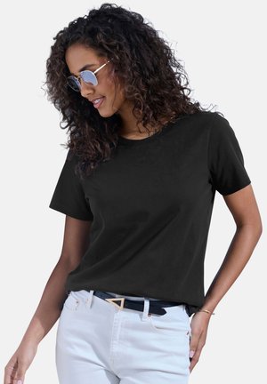 Frau mit lockigem dunklem Haar, die eine Sonnenbrille, ein schwarzes T-Shirt mit kurzen Ärmeln, weiße Hose und einen schwarzen Gürtel mit dreieckiger Schnalle trägt.