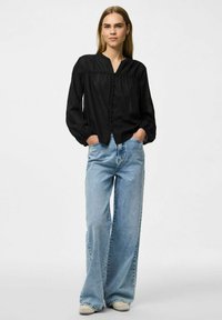 Svart blus med puffärmar och V-ringning, knappstängning och samlad detalj, kombinerad med ljusblå vida jeans och beige skor.