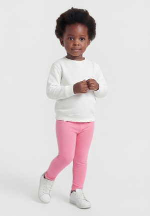 Jeune enfant debout, portant un t-shirt blanc à manches longues, des leggings roses et des baskets blanches sur un fond clair uni.