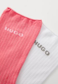 Dva páry ponožiek: jedny ružové s vertikálnymi pruhmi a bielym logom "HUGO", jedny biele s tenkými pruhmi a sivým logom "HUGO".
