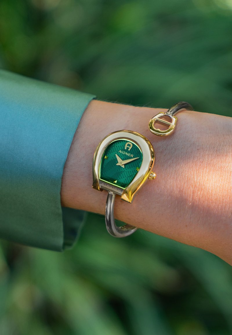 Orologio dorato con quadrante verde testurizzato, caratterizzato da una forma unica a ferro di cavallo e dettagli in metallo argento, indossato su un polso.