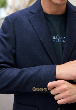 Mornarica sako izrađena od teksturiranog materijala, s rebrastim reverima i četiri smeđa dugmeta na manžetama. Nosi se preko tamne košulje s plavim tekstom.