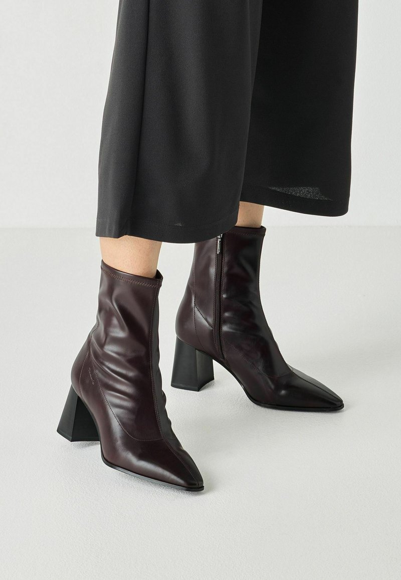 Γυναίκες που φορούν σκούρα καφέ ankle boots με ψηλό τακούνι και πλευρικά φερμουάρ, μαζί με μαύρο παντελόνι φαρδιάς γραμμής, σε απλό ανοιχτό φόντο.