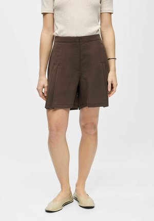 Vrouw die donkerbruine knielange shorts, beige korte mouwen top en beige platte schoenen draagt, staand tegen een effen lichte achtergrond.
