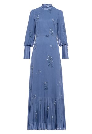 Nainen pukeutuneena pitkähihainen, korkeakauluksinen sininen mekko, jossa on kukkakuosi ja laskostettu helma, seisoo käsi lanteilla.