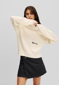 Übergroßer, cremefarbener Strickpullover mit gerippten Bündchen und Saum, versehen mit einem schwarzen Logo-Detail, kombiniert mit einem schwarzen Denim-Minirock.