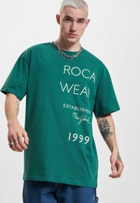Grøn bomulds t-shirt med afslappet pasform, korte ærmer og hvid trykt tekst: "ROCA WEAR ESTABLISHED New York 1999."