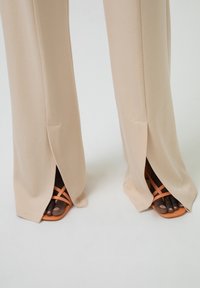 Pantaloni bej cu picioare largi și fante laterale, asortați cu sandale cu bretele portocalii. Materialul pare neted, având un design minimalist.