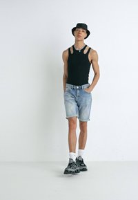 Débardeur noir, shorts en denim bleu clair, chapeau seau noir et baskets à motifs. Le modèle se tient les mains dans les poches devant un fond blanc.