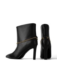 Bocanci din piele neagră până la gleznă, cu vârfuri ascuțite, tocuri înalte și un accent din lanț de aur în jurul gleznei. Textură netedă și formă elegantă.