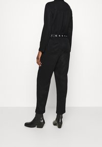 Combinaison noire avec manches longues, taille ceinturée avec des détails en logo, et jambes fuselées, associée à des bottines noires à talons.