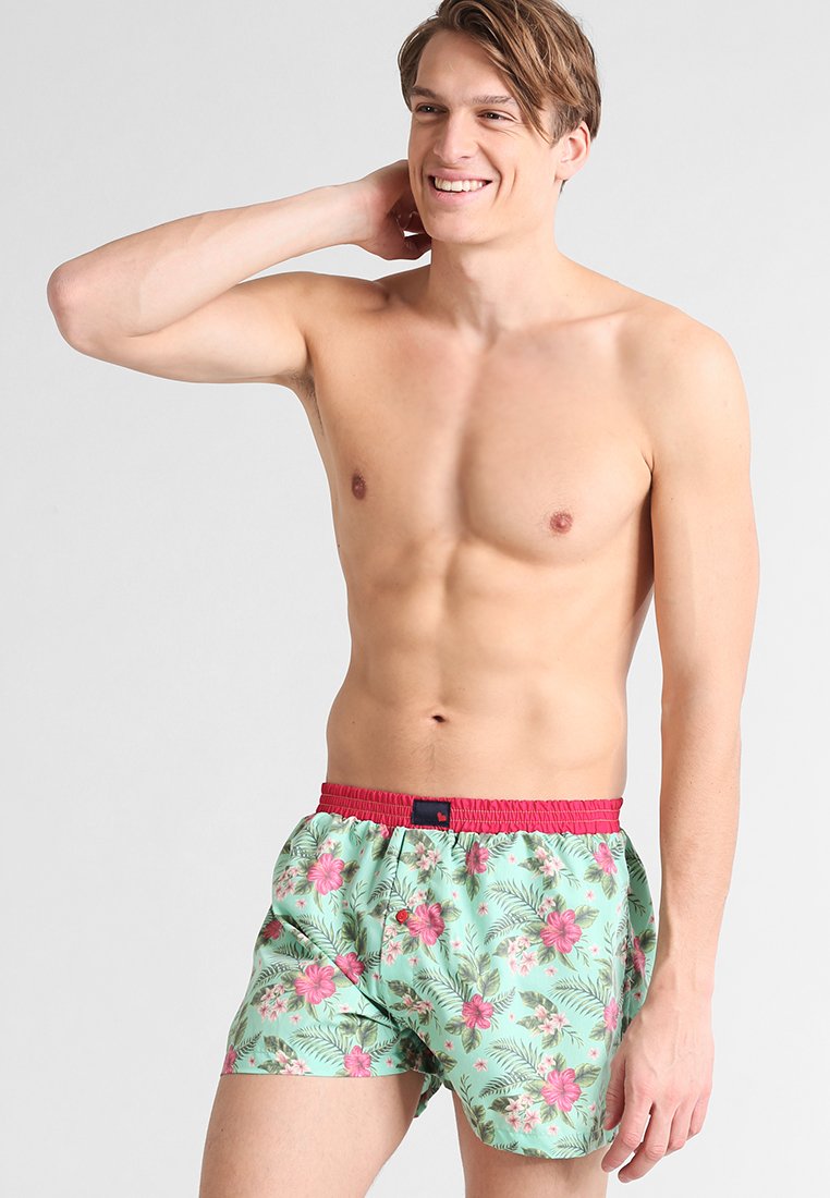 Costume da bagno verde con motivi floreali rosa tropicali, elastico in vita rosso e dettaglio con bottone sul davanti.