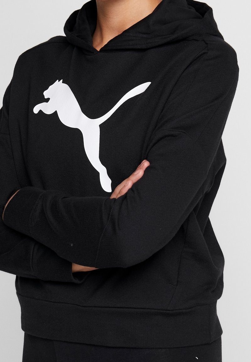 Personne portant un sweat à capuche noir avec le logo Puma blanc sur la poitrine, bras croisés, sur un fond gris clair.