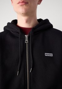 Černá mikina na zip vyrobená z měkké látky, s kovovým zipem a malou štítkovou nášivkou. Kapuce s tkaničkami pro úpravu.