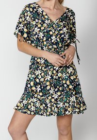 Žena oblečená v krátkých květinových šatech s volánkovým lemem a zavazovacími rukávy, které mají žluté, bílé a zelené květy na černém pozadí.