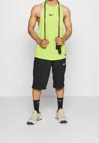 Persona musculosa con una camiseta sin mangas de neón y pantalones cortos negros sostiene las empuñaduras de una cuerda para saltar negra, de pie sobre un suelo gris contra una pared blanca.