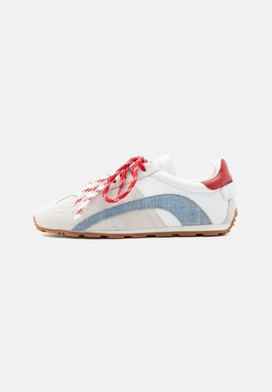 Sneaker bianca e beige con striscia curva in denim blu, linguetta rossa sul tallone, lacci rossi e bianchi e suola in gomma, vista laterale su sfondo bianco.