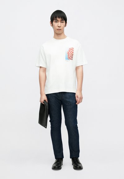 T-shirt blanc en coton avec un motif de parapluie rouge et bleu, associé à un jean bleu foncé et des chaussures noires, tenu avec une pochette sombre.