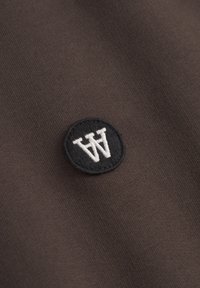 Bruine stof met een gladde textuur, voorzien van een ronde, zwarte geborduurde patch met de letter "W" in het wit.