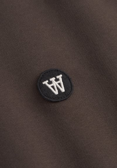 Tissu marron à la texture lisse présentant un patch rond noir brodé avec la lettre "W" en blanc.