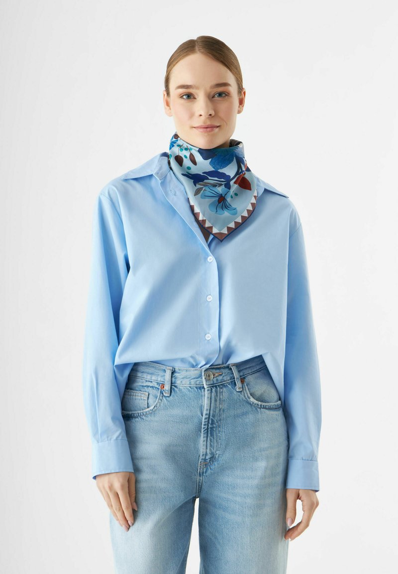 Chemise bleue à manches longues avec fermeture à boutons, associée à un jean bleu clair. L'écharpe présente des motifs floraux dans des teintes de bleu, rouge et marron.