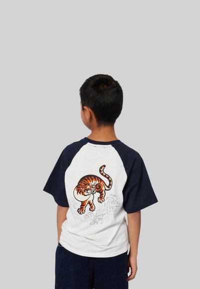 T-shirt à manches courtes avec un corps blanc et des manches raglan navy. Il présente un graphique d'un tigre orange au dos avec des accents de vagues.