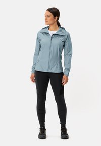 Lichtblauwe, lichte zip-up jas met capuchon, met een soepele textuur en subtiele branding, gecombineerd met zwarte strakke leggings en wandelschoenen.