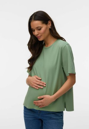 Gravid kvinde med langt mørkt hår iført grøn kortærmet skjorte og blå jeans, der blidt holder om maven og smiler med lukkede øjne.