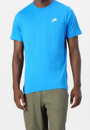 Mężczyzna w jaskrawoniebieskiej krótkiej koszulce Nike i oliwkowych spodniach, stojący na białym tle.