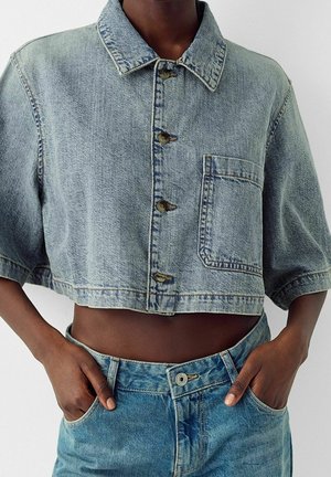 Osoba o ciemnej karnacji ubrana w krótki jasnoniebieski dżinsowy zapinany na guziki płaszcz i wysokowodne niebieskie dżinsy z rękami w kieszeniach.