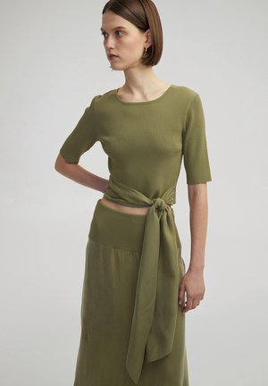 Olijfgroene, geribbelde gebreide top met korte mouwen, geknoopt in de taille. Gecombineerd met een bijpassende rok met een gladde textuur en een brede tailleband.