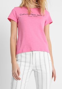 Růžové bavlněné tričko s krátkým rukávem, se černým potiskem "Calvin Klein Jeans od roku 1978". Kombinováno s bílými pruhovanými kalhotami.