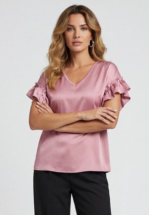 Femme aux cheveux blonds ondulés portant un chemisier en satin rose avec des manches courtes volantées et un pantalon noir, bras croisés, fond neutre.