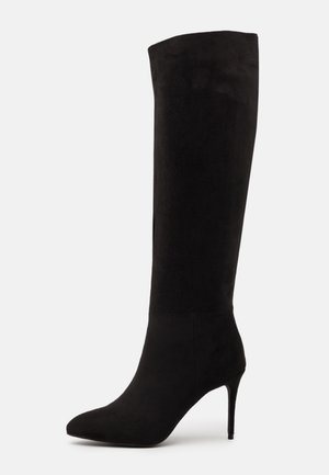 Bota de ante negra hasta la rodilla con tacón de aguja alto y puntera puntiaguda sobre un fondo blanco.