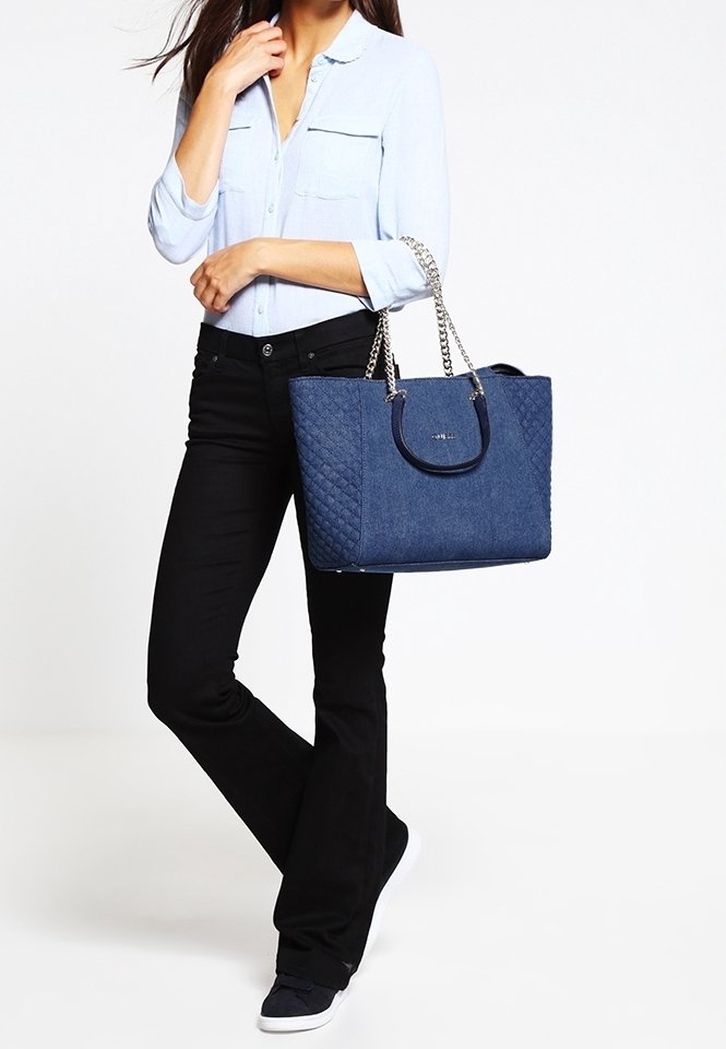 Femme portant un chemisier bleu clair et un pantalon noir, tenant un grand sac à main matelassé bleu avec des poignées en chaîne sur le côté.
