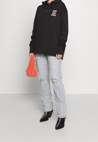 Osoba v černé mikině s růžovým květinovým logem, roztrhaných světle modrých džínách, černých špičatých botách, držící malou načechranou oranžovou tašku.