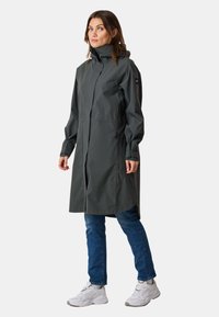 Cappotto impermeabile grigio scuro con colletto alto e cappuccio, dotato di cerniera anteriore e tasche laterali; abbinato a jeans blu e sneaker bianche.