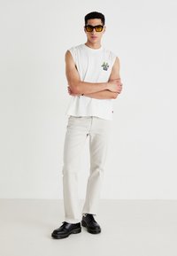 Witte mouwloze katoenen shirt met een bloemendessin, gecombineerd met lichtbeige broek en zwarte veterschoenen met een dikke zool.