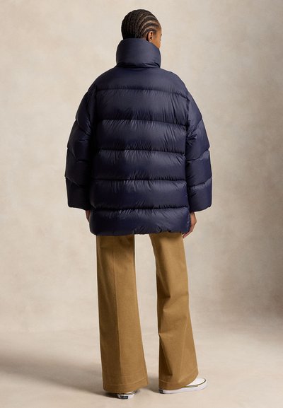 Personne aux cheveux tressés portant une doudoune bleu marine, un pantalon large marron et des baskets blanches, debout devant un fond neutre.