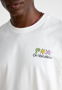 Ingrandimento di una persona che indossa una maglietta bianca con un piccolo palmo, sole, stella di mare e la scritta "In Vacanza" sul petto.