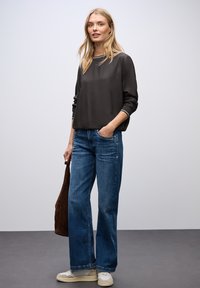 Top lungo in raso nero con scollatura rotonda, abbinato a jeans in denim blu a gamba larga. Il modello tiene una borsa in suede marrone e indossa sneakers color crema.