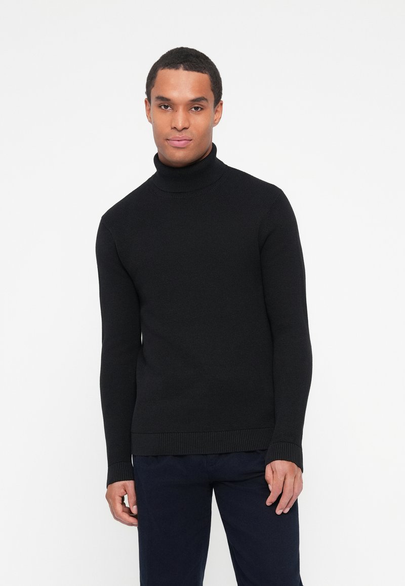 Maglione nero a collo alto in tessuto a coste con maniche lunghe e design aderente. Indossato con pantaloni blu scuro, su uno sfondo bianco uniforme.