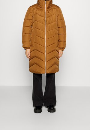 Długi brązowy pikowany płaszcz z przednim zamkiem błyskawicznym, wysokim kołnierzem, bocznymi kieszeniami, noszony na czarnych rozszerzanych spodniach i czarnych masywnych butach.