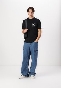 Μαύρο t-shirt με στρογγυλή λαιμόκοψη, διακοσμημένο με γραφικό σχέδιο, σε συνδυασμό με ανοιχτό μπλε cargo jeans με πολλαπλές τσέπες και λευκά sneakers.