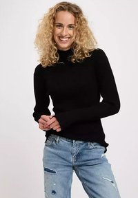 Schwarzer, langärmliger, gerippter Pullover mit Stehkragen, kombiniert mit hellblauen, distressed Jeans. Der Pullover hat eine figurbetonte Silhouette.