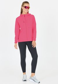 Rosa lättjacka med huva, dragkedja fram och två bröstfickor. Kombinerad med svarta leggings och grå träningsskor. Reflexiva solglasögon.