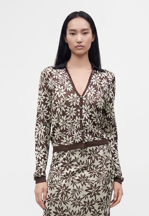Femme aux longs cheveux noirs portant un cardigan boutonné à motif floral marron et blanc et une jupe assortie sur un fond uni.
