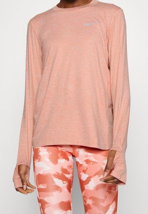 Vrouw draagt een perzikkleurig langemouwen sportshirt met duimgaten en bijpassende leggings met een koraalwit patroon.