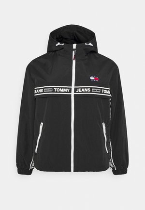 Musta huppari-tuulitakki, jossa valkoiset vetoketjut, joustavat hihansuut ja vaakasuuntainen "Tommy Jeans" -brändäys rinnassa ja hihoissa.