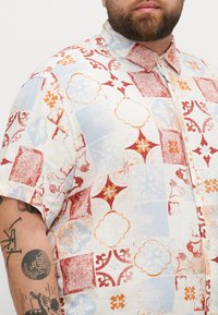 Chemise à manches courtes blanche avec des motifs en patchwork rouge, bleu et orange, présentant des motifs floraux et géométriques, fabriquée à partir d'un tissu léger.